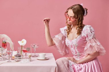 Günaydın kraliyet kahvaltısı. Çekici sarışın prenses portresi, süslü barok bir elbise giymiş uzanmış masada oturuyor ve pembe stüdyo arka planında kahve hazırlıyor. Ortaçağ, güzellik, yiyecek kavramı