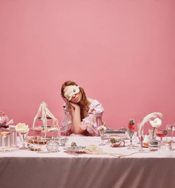 Rüya gören genç prenses. Masada oturan ve pembe stüdyo arka planında uyuyan elbise ve uyku maskesi takan sevimli kraliçenin portresi. Ortaçağ, güzellik, moda, ruh hali, günlük rutin kavramı