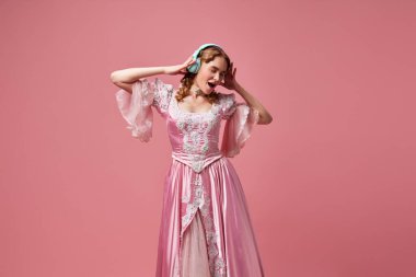 Modern prenses. Şirin sarışın prenses, süslü elbiseler giyen ve pembe stüdyo arka planında müzik dinleyen kraliçe. Alet kavramı, ortaçağ, güzellik, eski moda kıyafetler, reklam.