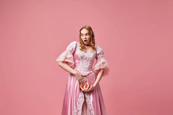 Femininity. Portrait of young beautiful woman, queen wearing dress and holding grapefruit down with surprised face over pink studio background. Concept of female rights, medieval, beauty, food