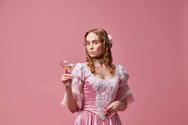 Elegant lady with cocktail. Shot of beautiful princess, queen wearing renaissance pink dress and standing with wine glass on studio background. Concept of medieval, beauty, party, alcohol, celebration