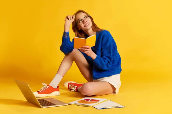 Exams preparations. Portrait of young pretty girl, student wearing eyeglasses sitting on floor surrounding books and laptop over yellow studio background. Concept of education, knowledges, university