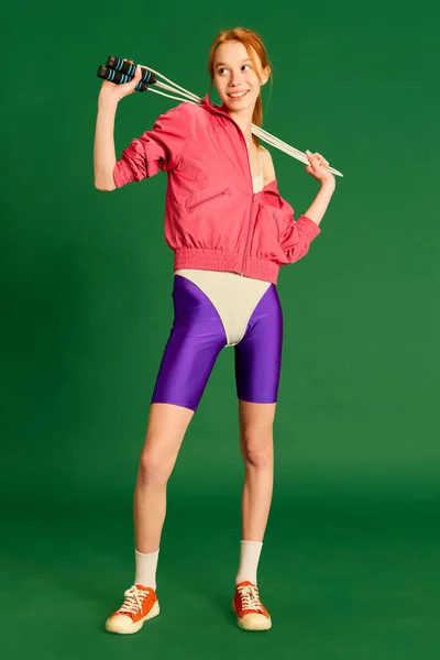 ワークアウトをジャンプ 緑のスタジオの背景に笑顔でロープジャンプでスポーツウェアの訓練を身に着けている陽気な赤い髪の若い女の子 スポーツ フィットネス 健康的なライフスタイル ファッション アクティビティの概念 — ストック写真