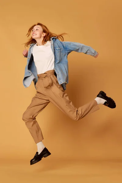 Happiness, joyful. One young red haired pretty girl, student wearing casual clothes jumping with joy at camera over ginger background. Youth, beauty, positive emotions, sales, ad concept