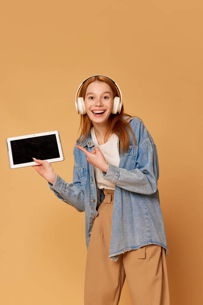 Онлайн школа. Портрет с ученицей, девушка в наушниках, держащая планшет и указывающая на пустой экран с улыбкой на имбирном фоне. Образование, современные технологии, интернет, концепция эмоций