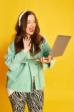 Çevrimiçi ders, ders. Genç bir öğrenci, kulaklık takan güzel bir kız dizüstü bilgisayar tutuyor ve sarı stüdyo geçmişine bakarak merhaba diyor. Eğitim, moda, yaşam tarzı, gençlik kavramı
