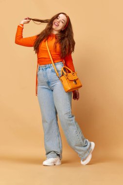 Neşeli öğrenci. Parlak kıyafetler giyen, kot pantolonlu, saçlarına dokunan ve kızıl arka planda gülümseyen genç, güzel bir kızın portresi. Moda kavramı, güzellik, yaşam tarzı, insan duyguları, reklam