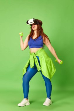VR gözlükleriyle antrenman. Genç, güzel bir kız, VR gözlüklü bir kadın dambıllarla egzersiz yapıyor ve yeşil arka planda kameraya gülümsüyor. Spor, fitness, sağlıklı, moda, aktivite kavramı