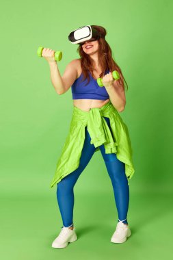 Üst beden eğitimi. Spor elbiseli ve video gözlüklü genç bir kız yeşil arka planda dambıllarla egzersiz yapıyor. Spor, fitness, sağlıklı, moda, aktivite kavramı