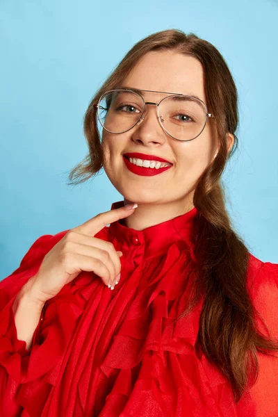 Happy, jolly life. Portrait of amazing woman in chic clothes with red lips touching chin and smiling at camera on blue studio background. Closeup. Concept of fashion, party, beauty, human emotions, ad