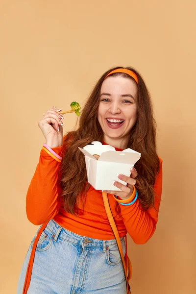 Healthy menu for a day. Portrait of pretty girl, student holding box with food and eating broccoli with smile on ginger background. Concept of diet, fitness, healthy food, sport, human emotions, ad
