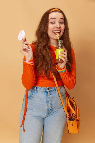 Cheat meal day. Portrait of happy, beautiful girl wearing bright clothes holding candy and drinking juice and smiling on ginger background. Concept of food, beauty, lifestyle, human emotions, ad