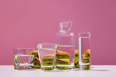 Fast food bağımlılığı. Pembe stüdyo arka planında su bardaklarının yanında bir sürü taze, lezzetli hamburgerle dolu bir hayat. Yiyecek kısıtlaması, diyet, yağ, karbonhidrat, abur cubur, sağlıksız gıda kavramı