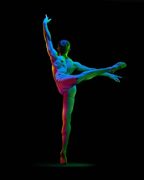 Portrait of handsome muscled male ballet dancer dancing over dark studio background with neon light. Back view. Weightless, flexible actor. Grace, art, beauty, contemporary dance concept