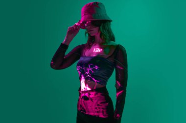Titreşim. Dijital neon ışıklı panama giyen genç bir kızın yaratıcı portresi. Koyu yeşil mod arka planda vücuda işlemeli filtre ışıkları. Dijital sanat, moda, siber punk, fütürizm kavramı