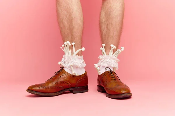 Funny male portrait with feet wearing classic shoes and white female socks with mushrooms inside over pink studio background. Concept of stylish clothes, food, beauty, art, creativity