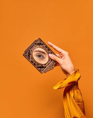 Görme. Turuncu arka planda kızların gözünün yansıması olan küçük bir ayna tutan bir kadın. Klasik moda kavramı, güzellik, sanat, yaratıcılık ve... İnsani duygular, yüz ifadesi