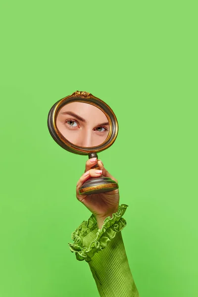 Expressive look. Female hand holding small mirror with reflection of charming womans face with natural make up on green background. Fashion, beauty, creativity, ad. Human emotions, expression concept