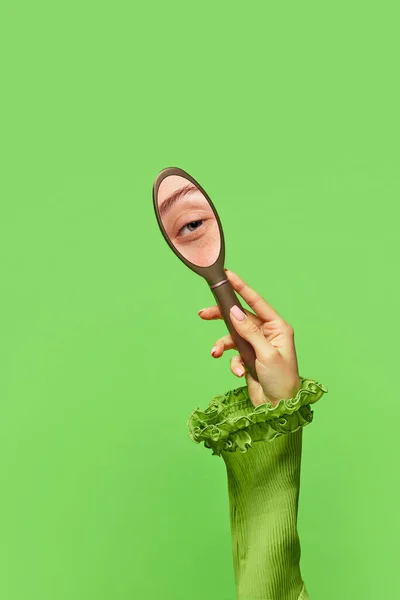 Sly female look. Portrait with womans hand holding oval little mirror with reflection of girls beautiful eye on green background. Beauty, art, creativity and ad. Human emotions, expression concept