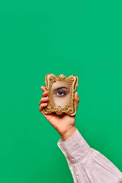 Female hand holding small mirror with reflection of charming womans eye with bright makeup over green background. Fashion, beauty, creativity, ad. Human emotions, expression concept