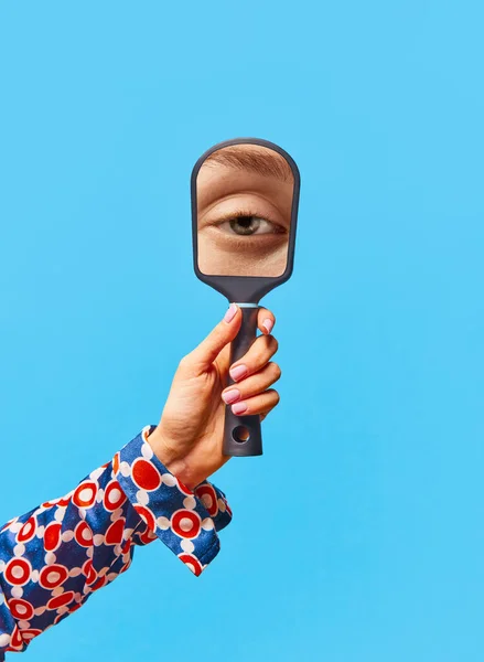 Vintage style. Female hand holding small mirror with reflection of girls eye without makeup over bright blue background. Concept of vintage fashion, beauty, art, creativity, human emotions, ad