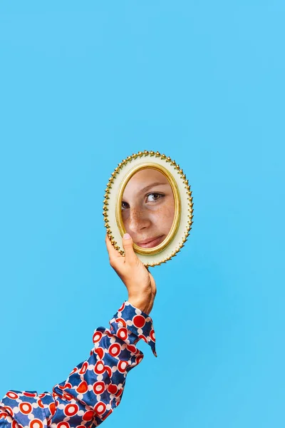 Female hand holding small mirror with reflection of beautiful, womans face with natural make up and freckles over blue background. Beauty, creativity, selfcare. Human emotions, expression concept
