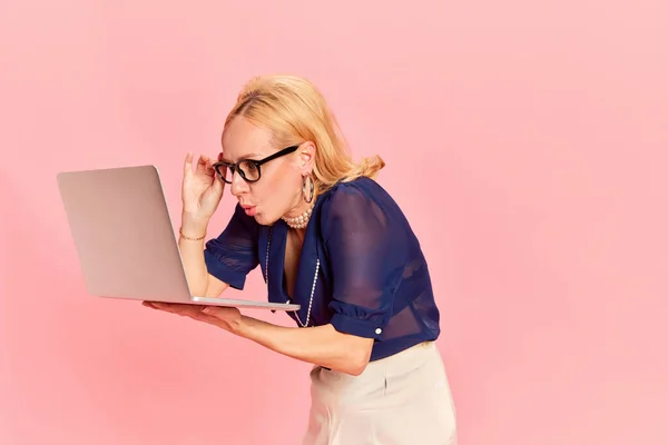 Business progress. Portrait of harming young woman, in retro clothes and glasses holding laptop with shocked face over pink background. Concept of emotions, career, beauty, fashion, vintage, retro, ad