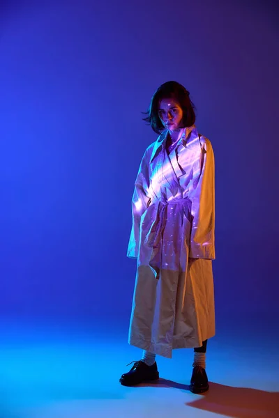 New fashion style. One attractive woman wearing trendy coat standing with neon reflection of space glow on body over blue mode background. Concept of digital art, fashion, cyberpunk, futurism