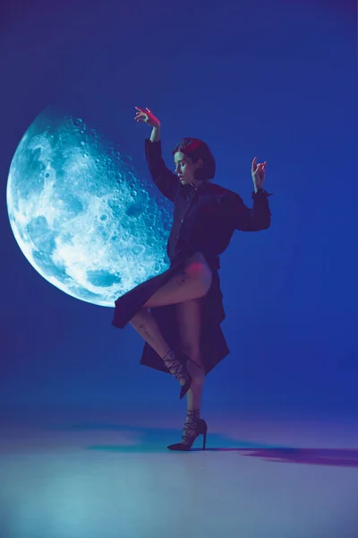 Full moon. One dreaming woman wearing elegant dress posing near projection over blue studio background in neon light. Concept of human emotions, digital art, fashion, futurism, art