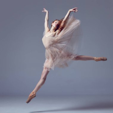 Esnekliğini göster. Genç ve inanılmaz derecede güzel bir balerin. Tüylü elbise giymiş. Gri stüdyo arka planında zarif bir şekilde zıplıyor. Güzellik konsepti klasik bale sanatı. Bale estetiği.