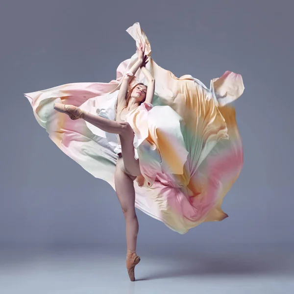 Aesthetic of classical dance. One adorable ballerina wearing rainbow dress emotional dancing with fabric over grey studio background. Concept of classic ballet, inspiration, beauty, dance, creativity