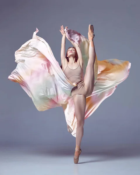 Aesthetic of classical dance. One grace ballerina wearing rainbow dress emotional dancing with fabric over grey studio background. Concept of classic ballet, inspiration, beauty, dance, creativity