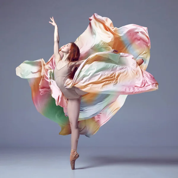 Tender soul. Young classical dancer wearing colorful flying dress dancing on fingertips over grey studio background. Concept of ballet, inspiration, beauty, contemporary dance, creativity, motion, ad