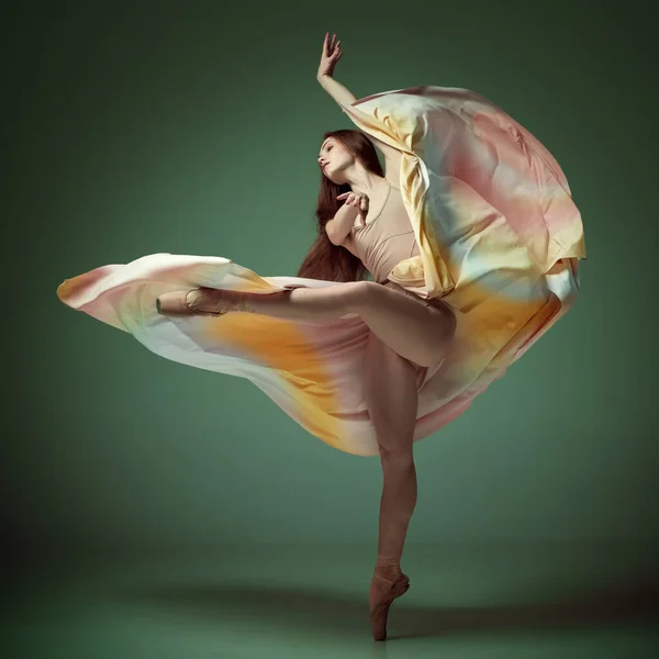 Tender soul. Young classical dancer wearing colorful flying dress dancing on fingertips over dark green background. Concept of ballet, inspiration, beauty, contemporary dance, creativity, motion, ad