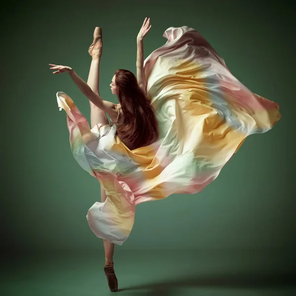 Modern ballet with silk dress. One young adorable professional ballet dancer wearing colorful dress dancing over dark green background. Concept of classical ballet, contemporary art, beauty, dance