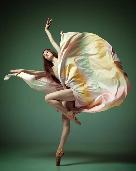 Modern ballet with silk dress. One adorable ballerina, young girl dancing gracefully with with fabric over dark green studio background. Concept of beauty classical ballet art, aesthetic