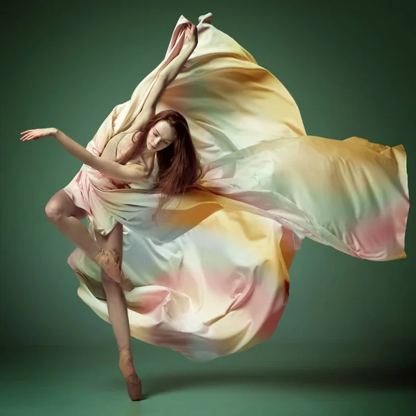 Portrait of inspired young ballerina, beautiful girl wearing silk dress and dancing with emotion over dark green background. Beauty of contemporary dance. Ballet art, action, flexibility, ad concept