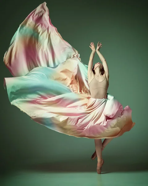Rainbow dress. Charming ballerina dancing elegant, grace movement with flying fabric over dark green studio background. Beauty of contemporary dance. Art, action, flexibility, inspiration, ad concept