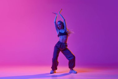 Genç, güzel, ince bir kız, hip-hop giysileri giyen dansçı. Gradyan mor neon arka planda dans ediyor. Çağdaş dans tarzı, ilham, hareket, hareket, hobi, moda, sanat, reklam kavramı