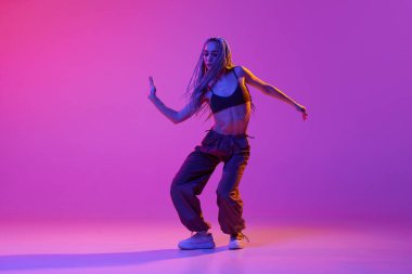 Hip-hop dans savaşı. Sokak tarzı kıyafetler giyen genç bir kız. Mor stüdyo arka planında neon ışıkta duygularıyla dans ediyor. Çağdaş tarz kavramı, hareket, sanat, hareket, ilham, reklam