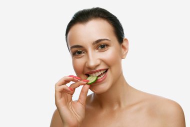 Alternatif ev güzelliği prosedürleri. Mükemmel cilt ısıran, salatalık dilimleyen ve beyaz arka planda gülümseyen güzel, genç bir kadının portresi. Güzellik, kaplıca, kozmetik, cilt bakımı, duygu konsepti