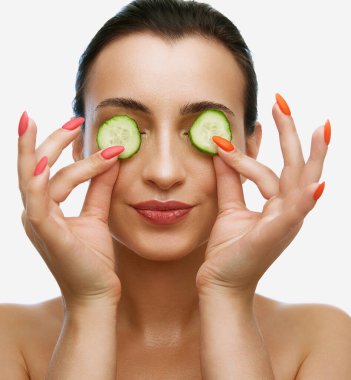 Güzel, esmer bir kadının portresi. Çıplak omuzları ve pürüzsüz bir teni var. Beyaz arka plandaki yamalar gibi salatalık dilimleri kullanıyor. Güzellik, kaplıca, kozmetik, cilt bakımı, sağlık, reklam konsepti