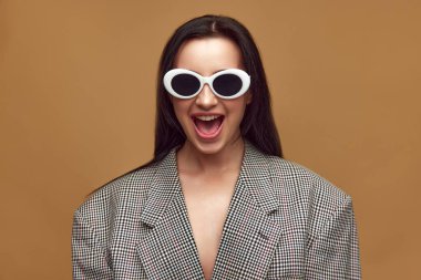 Parlak duygular. Güzel, şık bir kadının portresi, kahverengi stüdyo arka planında kareli ceket ve güneş gözlüğü takan bir kız. Güzellik kavramı, moda, trendler, cilt bakımı, insan duyguları, reklam