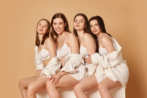 Girls pajama party. Cropped image with group of smiling woman with long hair sitting over beige studio background. Concept of natural beauty, body and skin care, healthy eating, fashion, cosmetics, ad