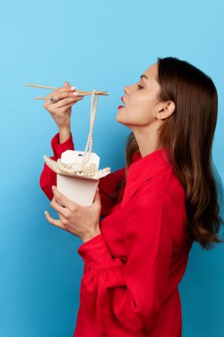 Çekici bir kadının portresi elinde Çin eriştesi çubukları ve mavi arka planda zevkle kutuda yemek yiyor. Çöp sanatı, parlak renkler. Yemek kavramı, güzellik, moda, fast food, satış, reklam