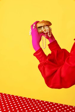 Sarı arka planda mücevherlerle dolu büyük bir burgeri tutan kadın ellerinin portresi. Çöp sanatı, parlak renkler. Yemek kavramı, mücevher, güzellik, moda, yemek, duygu, mutfak, yaratıcılık, reklam