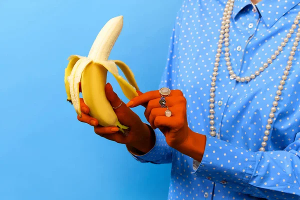 Portrait of colored female hands wearing rings holding banana over blue studio background. Trash pop art, bright colors. Concept of meal, jewelry, fruits, diet, nutrition, fashion, beauty, sales, ad