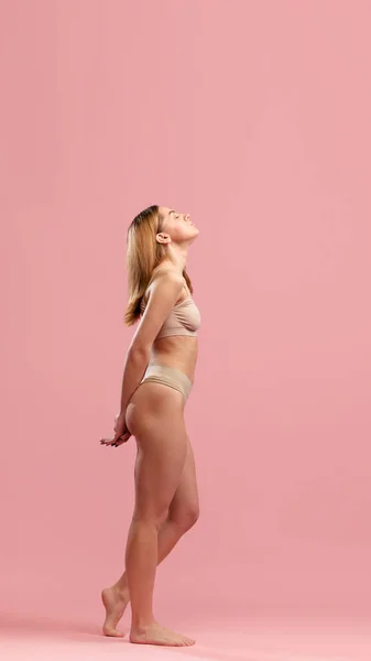 ピンクのスタジオの背景の上にポーズをとってベージュ色の下着を身に着けている若い夢の少女のサイドビューの肖像画 ファッション スキンケア ボディケア ウェルネス 広告コンセプト — ストック写真