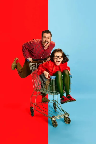 stock image Crazy family. Portrait with funny couple, man and attractive woman sitting at shopping cart and racing on blue background. Concept of fun, human emotions, love, relationship, adventures, sales