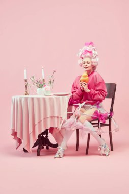 Portrait with dreaming princess, queen wearing big wig and starting eat huge croissant on pink background with astonished face. Concept of food, diet, comparison of eras, modernity and renaissance clipart